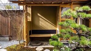 A traditional Japanese building with a wooden exterior, a tatami mat entrance, and a garden with pine trees and a stone path.