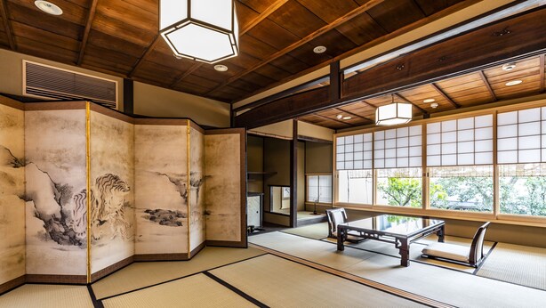 A traditional tatami mat room with a low table and benches, wooden beams on the ceiling, and a view of greenery outside.