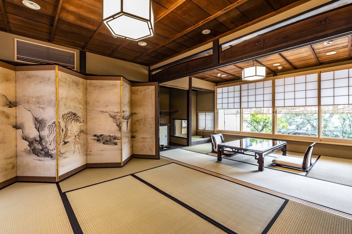 A traditional tatami mat room with a low table and benches, wooden beams on the ceiling, and a view of greenery outside.