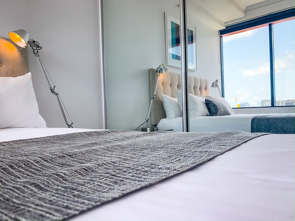 Quarto de hotel moderno com uma cama grande, luminária de mesa, cabeceira estofada e vista para a cidade através de amplas janelas.