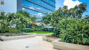 A modern outdoor area with a tiled floor, surrounded by lush greenery and a large glass building in the background. - Apartments on Connor (Fortitude Valley)