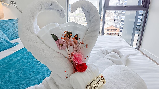 A bedroom with a large window, a bed with white bedding, a blue pillow, and a decorative swan sculpture with flowers. - Apartments on Connor (Fortitude Valley)