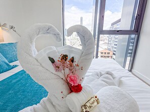 Quarto com uma janela grande, cama com roupa de cama branca, travesseiro azul e uma escultura decorativa de cisne com flores.