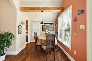 A dining area with a wooden ceiling, a round table, chairs, a hanging light fixture, and a window with blinds.