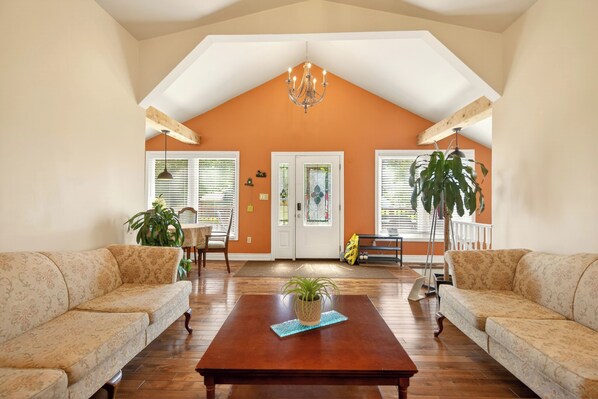 A spacious living room with a high ceiling, wooden beams, a chandelier, two sofas, a coffee table, and a door with glass panels.