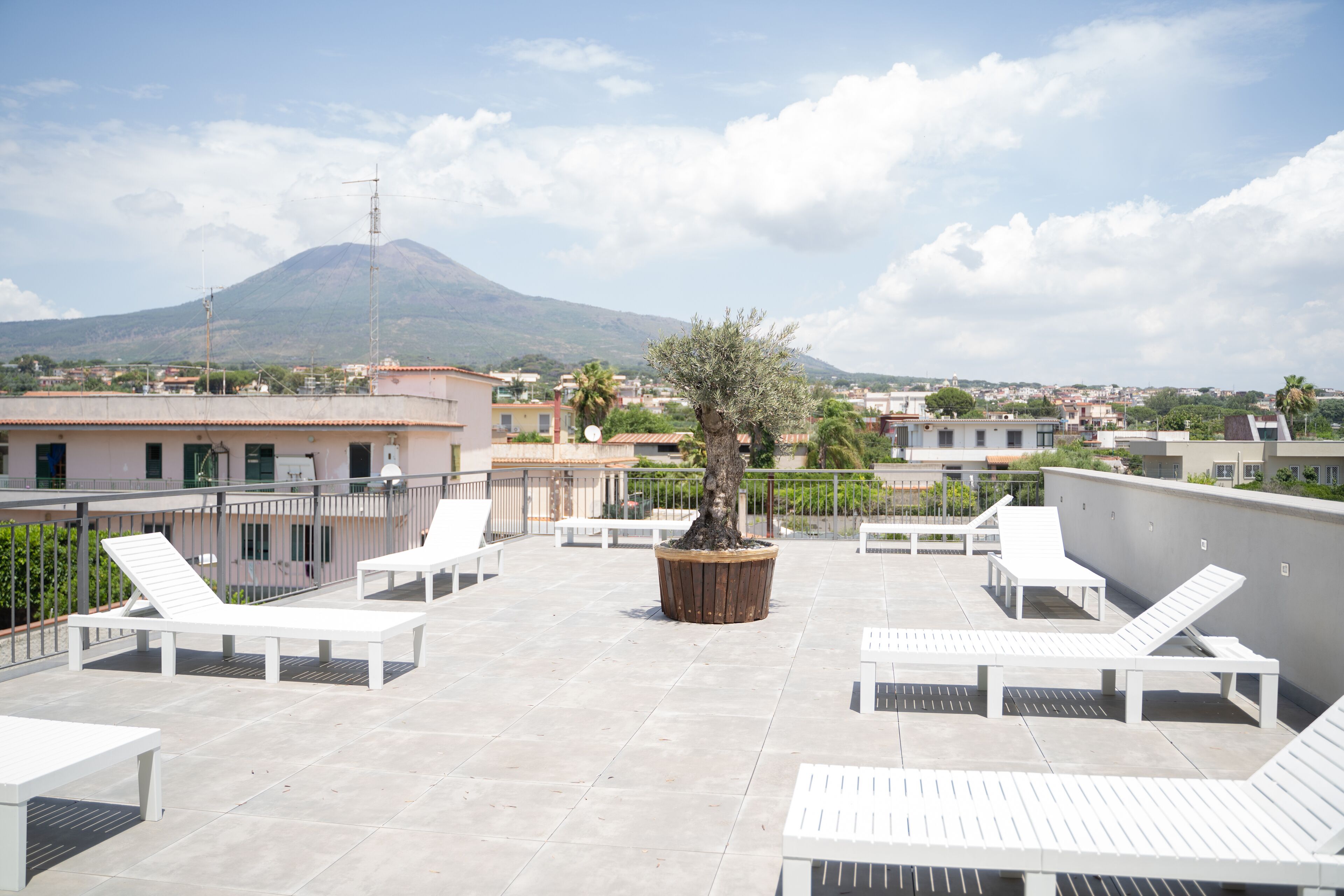 Eine Dachterrasse mit weißen Liegestühlen, einem Blumentopfbaum und Blick auf einen Berg.
