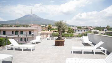 Eine Dachterrasse mit weißen Liegestühlen, einem Blumentopfbaum und Blick auf einen Berg.