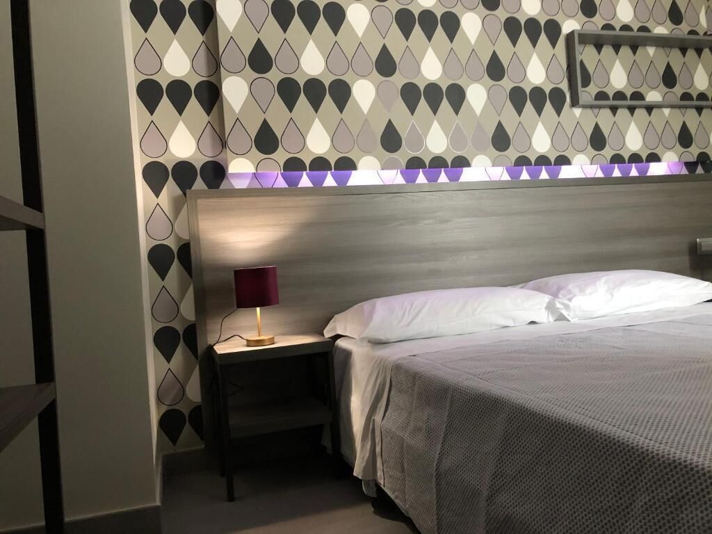 A neatly made bed with a grey bedspread and white pillows in a hotel room with a patterned wallpaper and a wooden headboard.