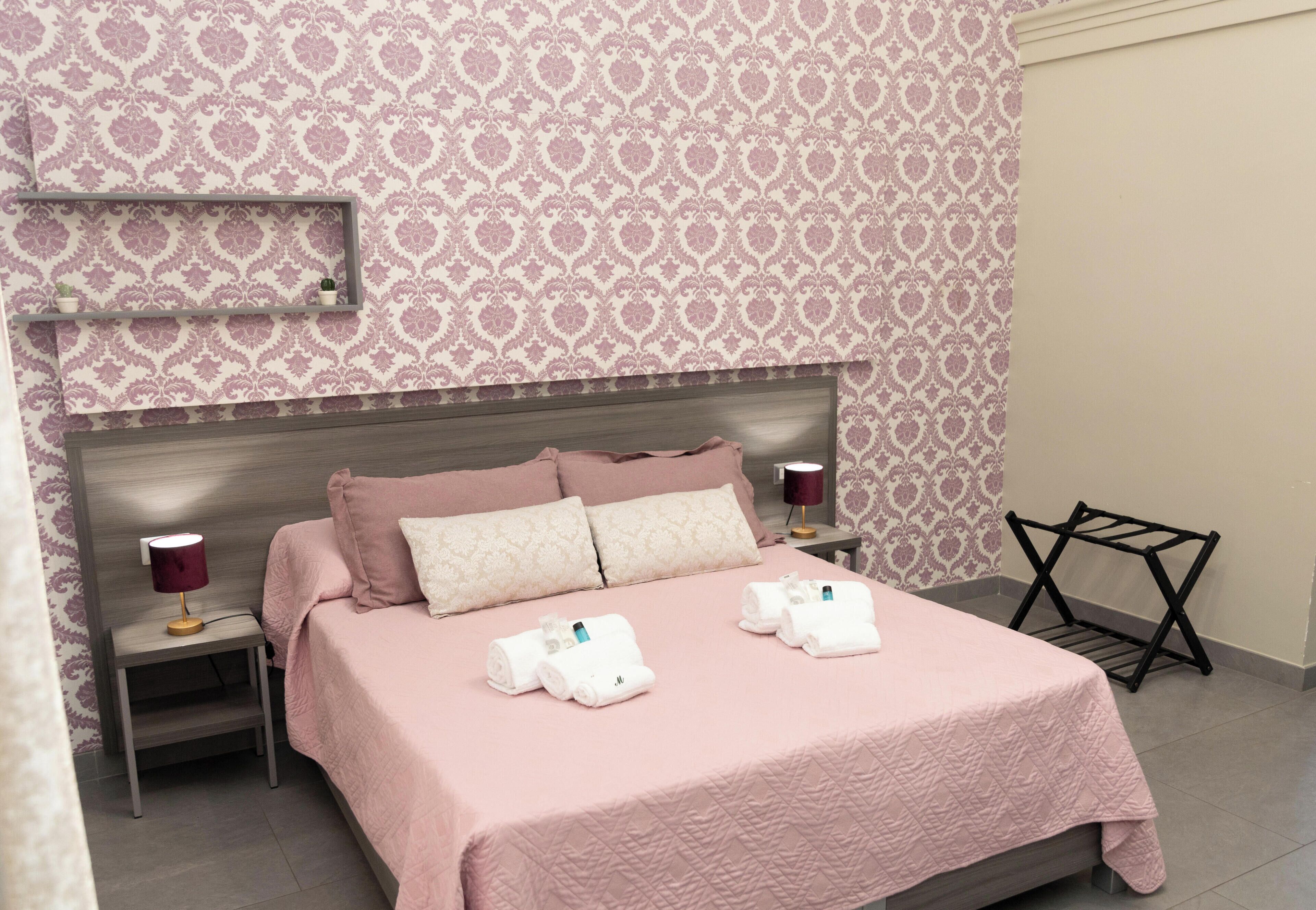 Una cama bien hecha con una colcha rosa, dos almohadas y dos toallas blancas. Hay dos mesitas de noche con lámparas, un estante en la pared con objetos decorativos y un tendedero negro.