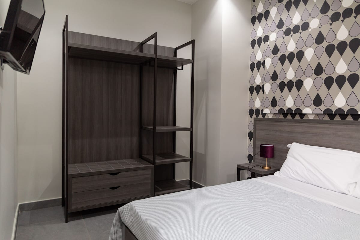Um quarto com cama, guarda-roupa com prateleiras abertas, mesa de cabeceira com abajur e parede estampada.