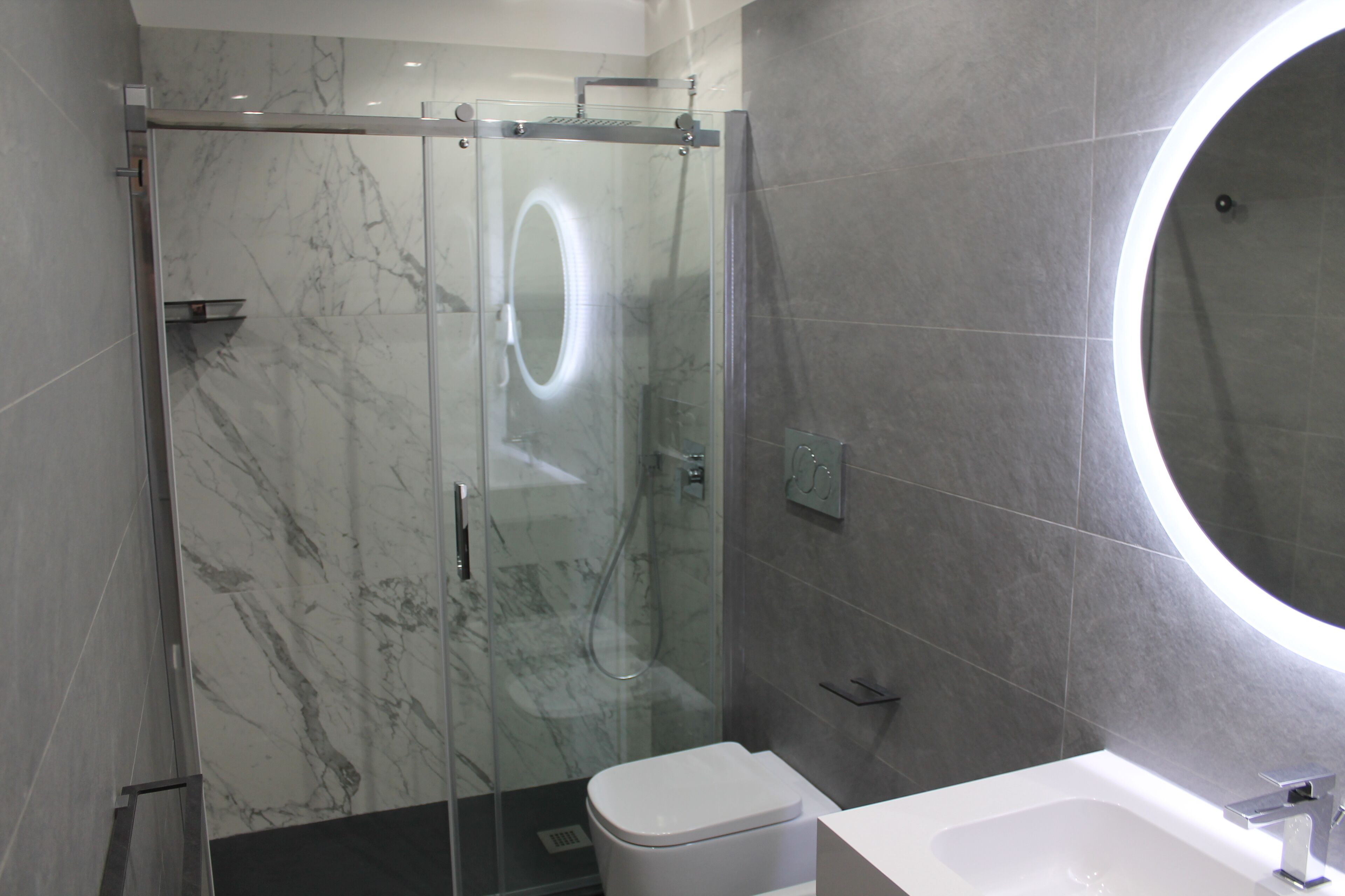 Un baño moderno con ducha envuelta en vidrio, inodoro blanco y un espejo grande con marco circular.