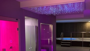 Un moderno espacio de spa con un jacuzzi, una sauna con paredes de vidrio y una puerta iluminada en tonos rosados.