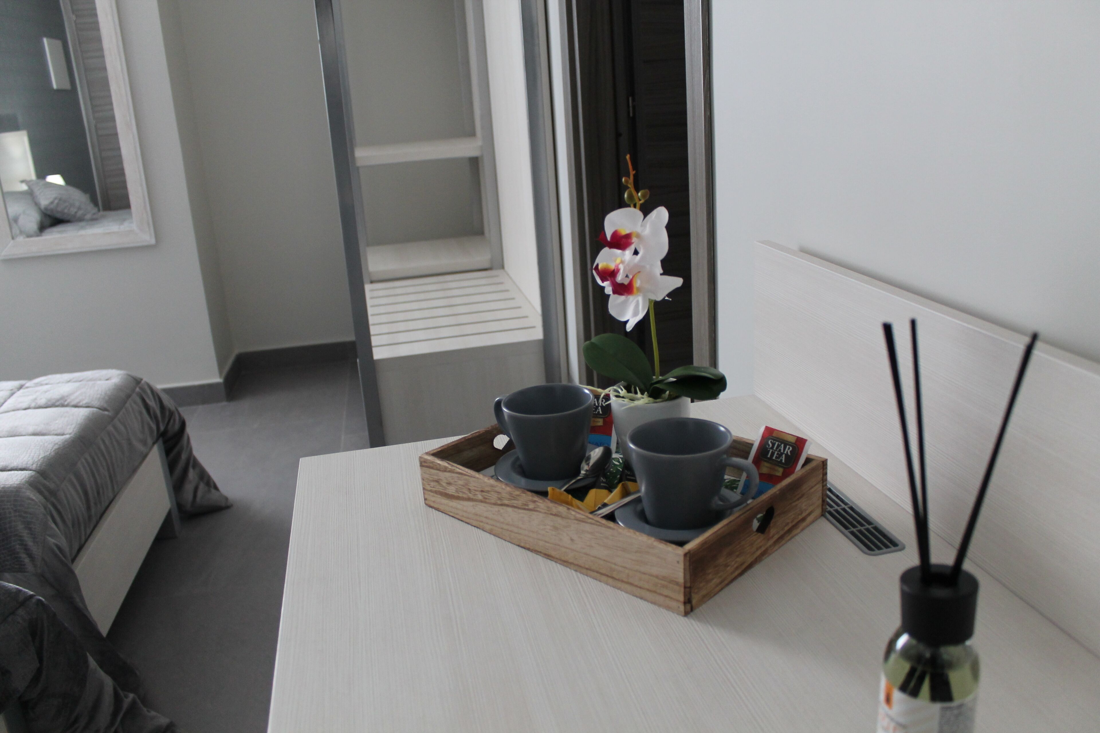 Habitación de hotel con una cama, una mesita de noche con una bandeja de tazas y una orquídea en maceta, y una ventana con vistas a un balcón.