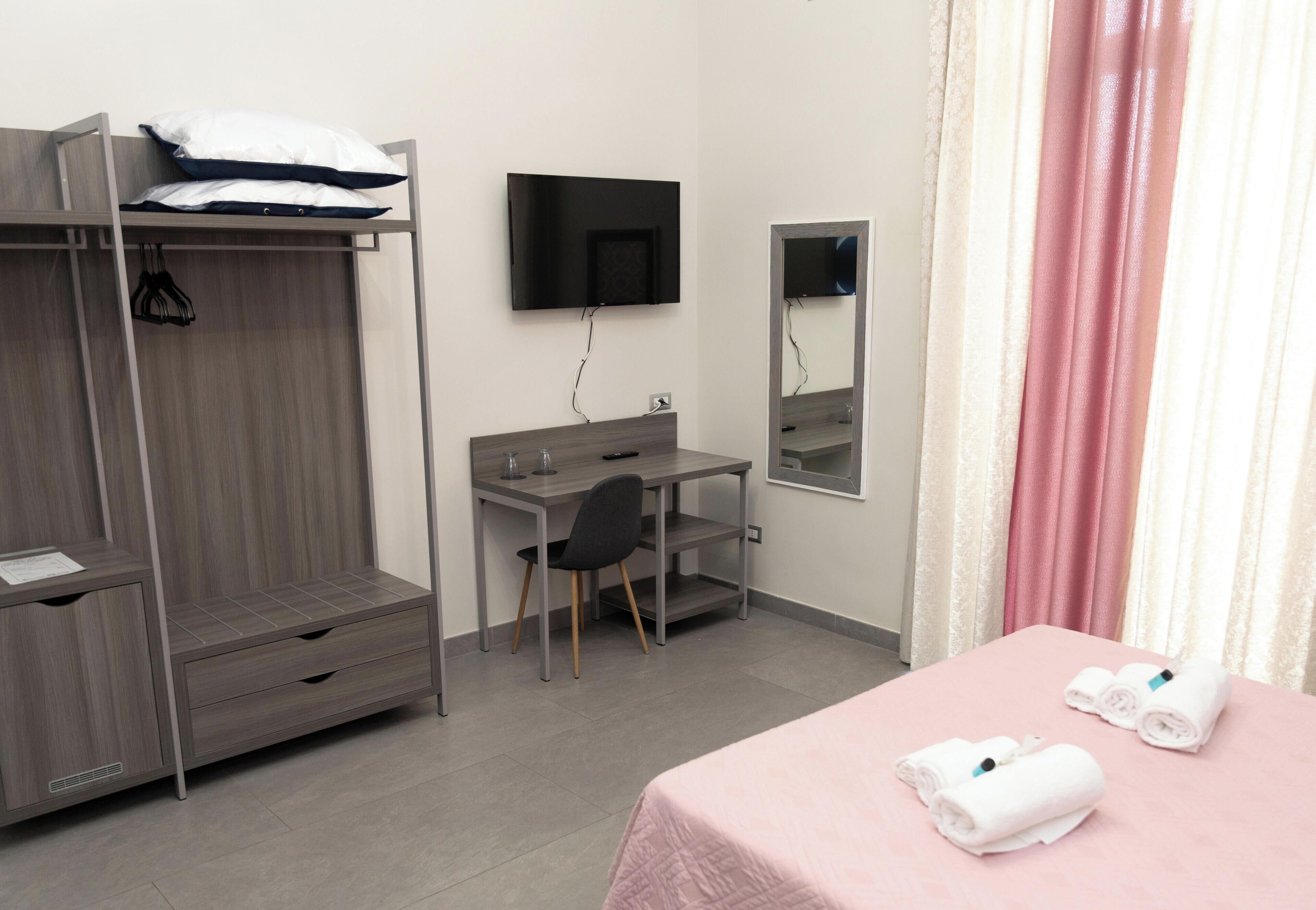 A hotel room with a bunk bed, a desk with a chair, a TV mounted on the wall, a mirror, and a bed with towels.