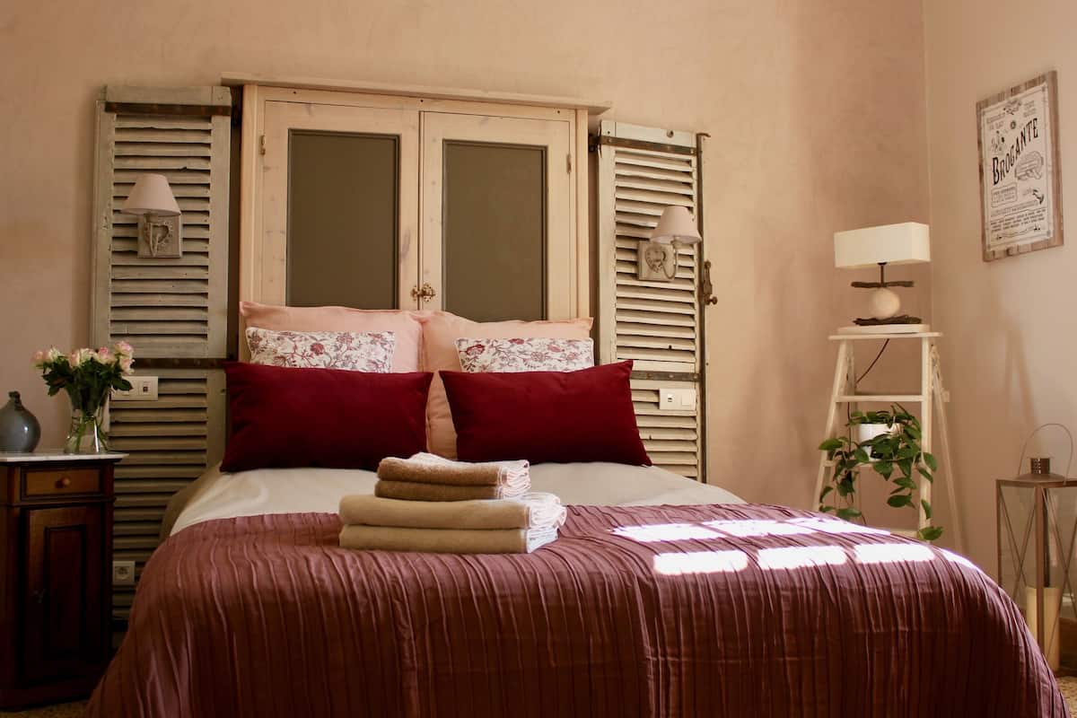 A bedroom with a bed, two windows with shutters, a bedside table with a lamp, and a wall-mounted picture.