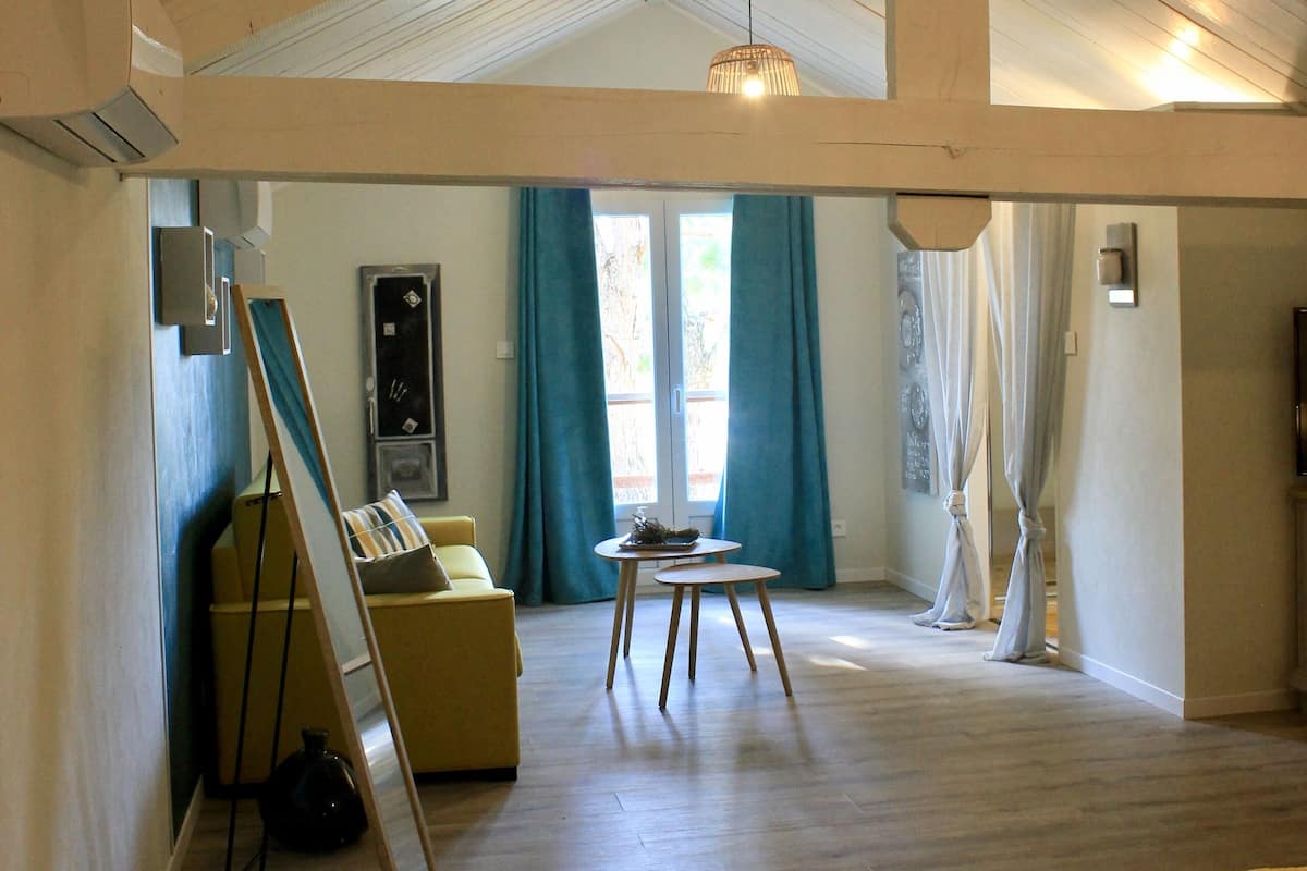 A spacious loft with a high ceiling, wooden beams, a large window with curtains, a sofa, a small table, and a ladder leaning against the wall.