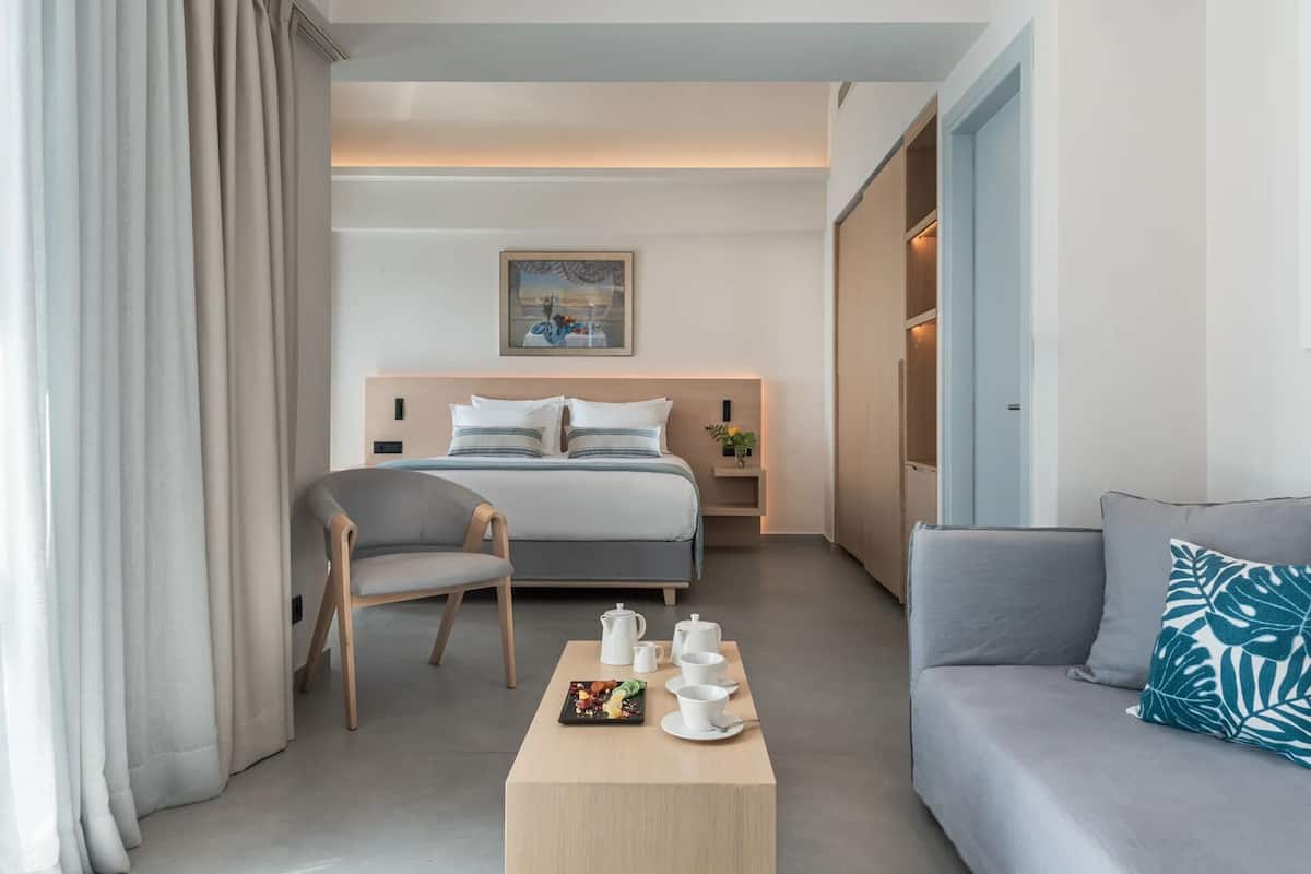 A modern hotel room with a bed, a gray sofa, a wooden coffee table with breakfast items, and a painting on the wall.