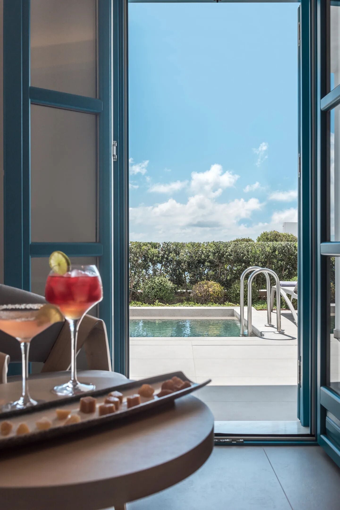 Vista de una piscina desde una puerta abierta, con una mesa al frente que presenta bebidas y aperitivos.