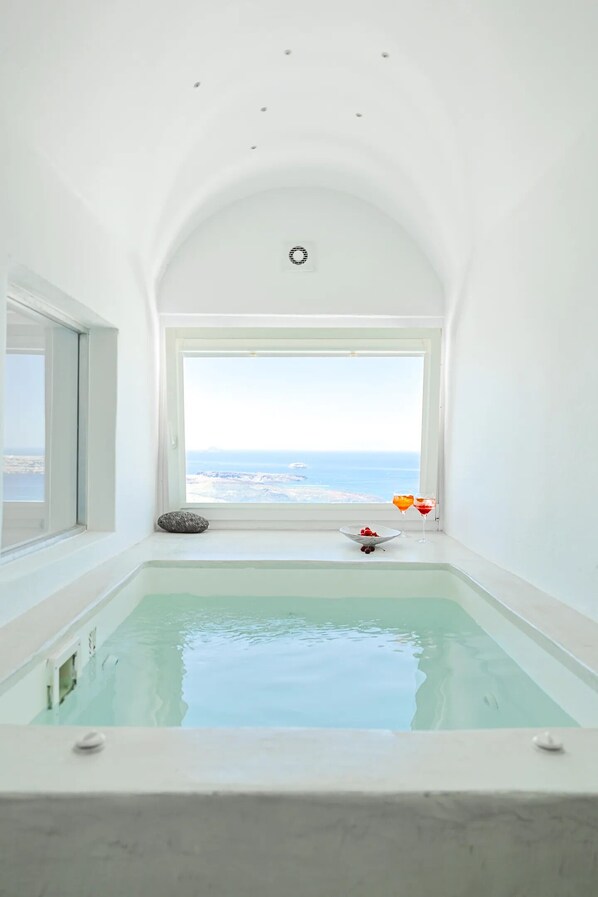 A spa room with a large window, a bathtub, and a bowl of fruit. - Aeifos Boutique Hotel (Santorini)