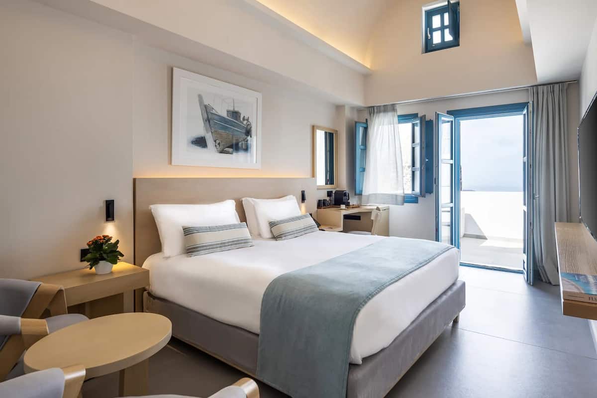 A modern hotel room with a large bed, a desk, a chair, a small table, and a balcony with a view.