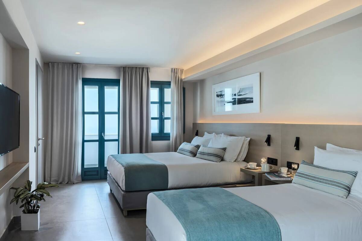 A hotel room with two beds, a TV, a window with curtains, a plant, and a framed picture on the wall.