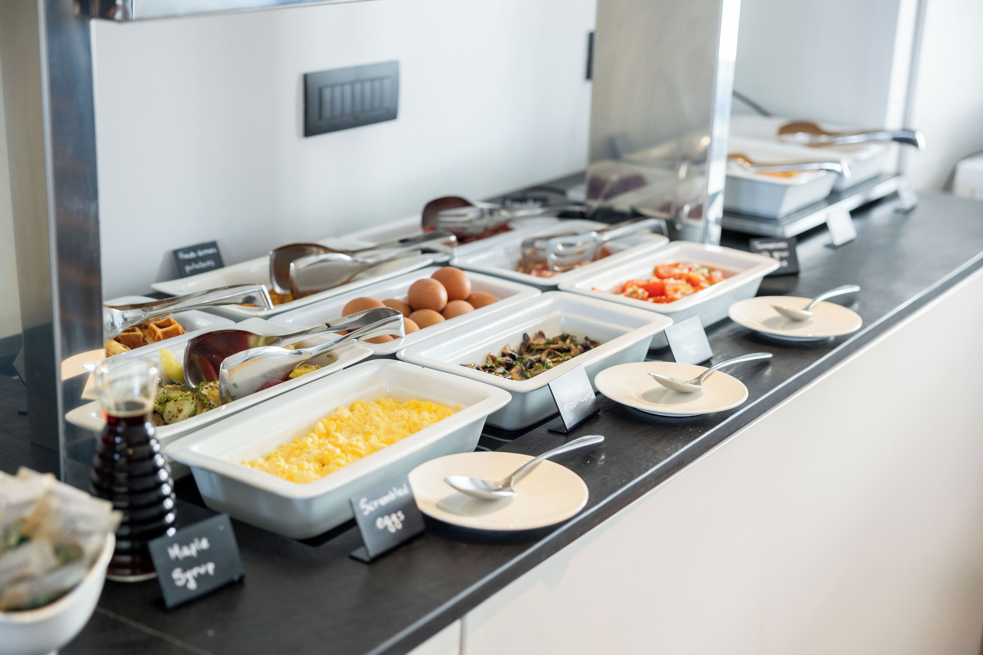 Un bufé con recipientes etiquetados de diversas comidas, incluyendo huevos revueltos, muffins y un dispensador de jarabe.