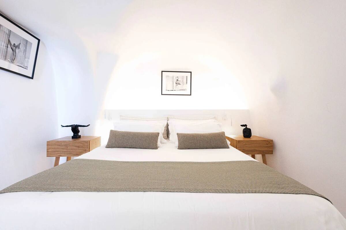 A neatly made bed with a striped bedspread, two pillows, and a headboard with a framed picture above it. There are two bedside tables with lamps and decorative items.