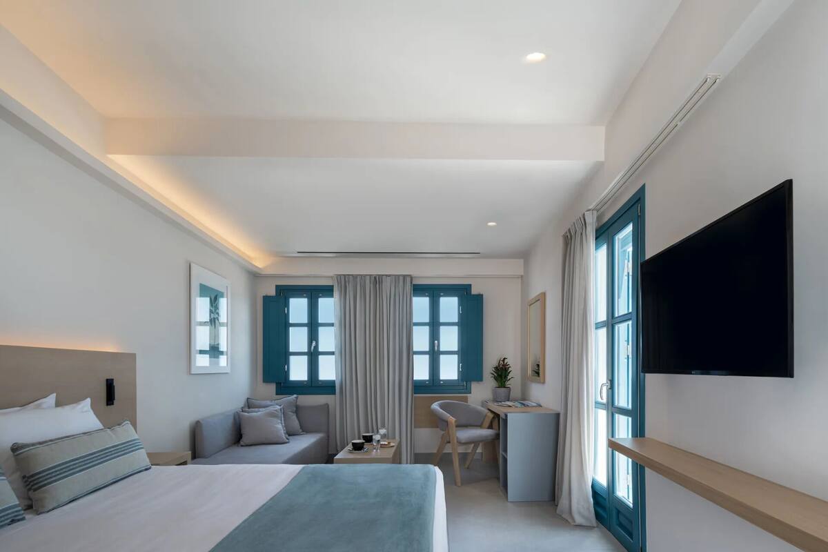 A modern hotel room with a bed, a desk, a chair, a TV, and large windows with curtains.