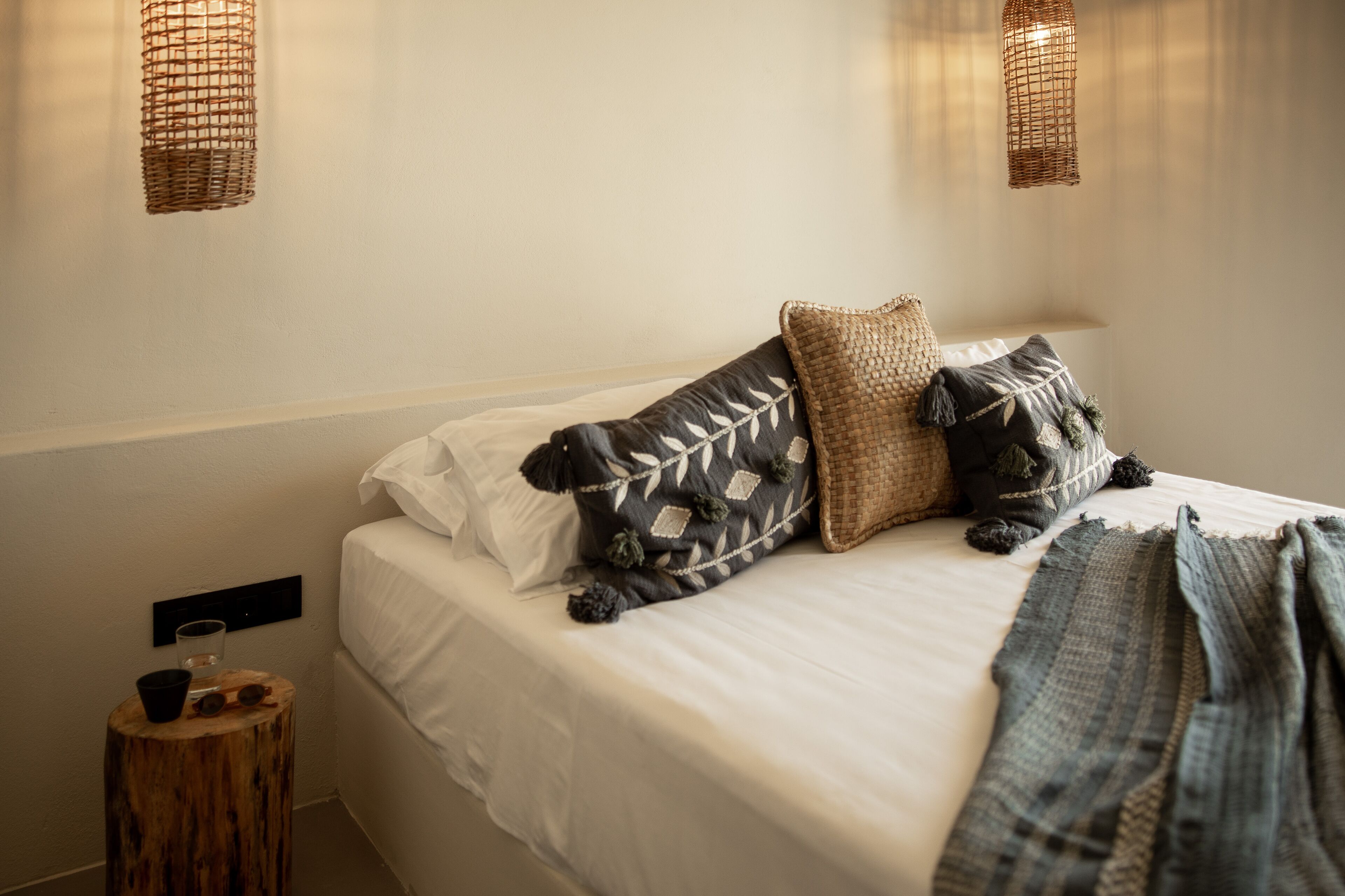 A neatly made bed with a white sheet, two decorative pillows, and a woven throw blanket. There is a small wooden stool with a glass and a cup beside the bed.