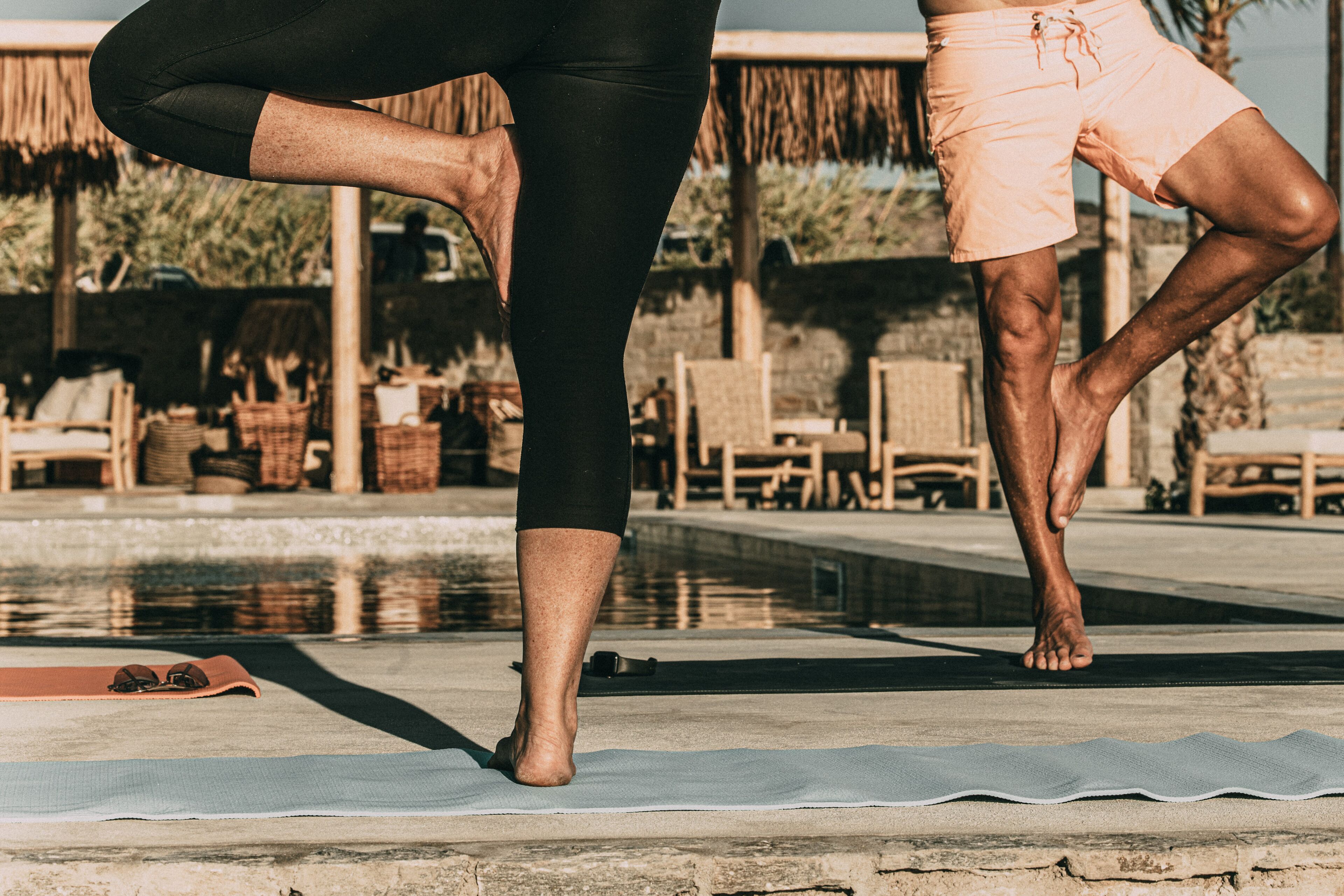 Een persoon doet een yoga-houding op een mat naast een zwembad, met ligbedden en een rieten dakconstructie op de achtergrond.