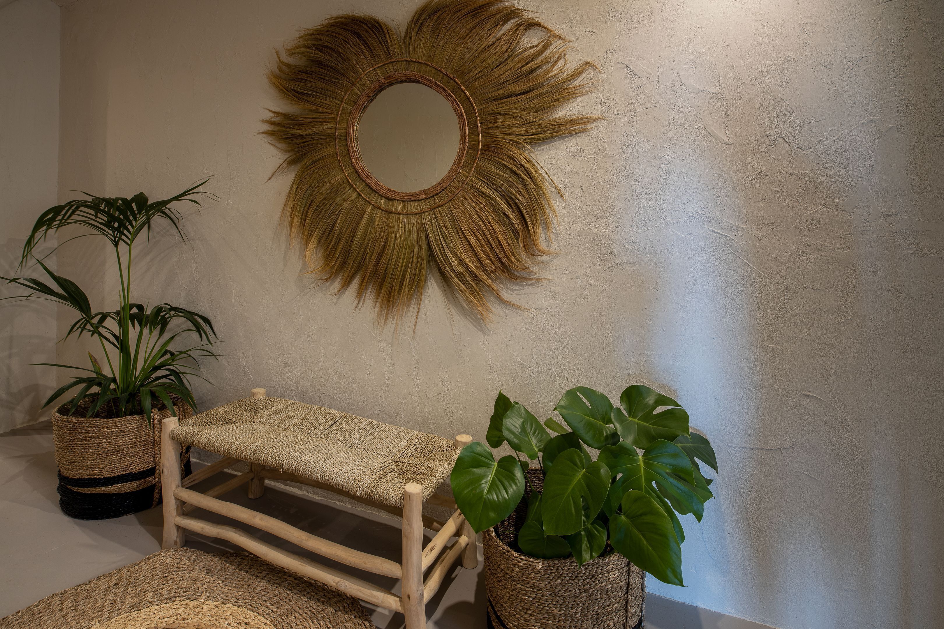 A room with a woven bench, two potted plants, and a sunburst mirror on the wall.