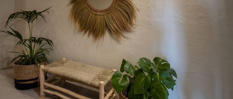 A room with a woven bench, two potted plants, and a sunburst mirror on the wall.