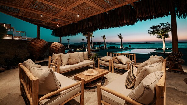 A cozy outdoor seating area with wooden furniture and a thatched roof, overlooking a pool and a sunset view. - Seesoo Paros Beachfront Luxury Hideaway (Paros)
