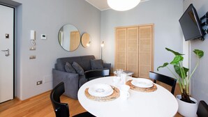 A modern living room with a round dining table set for two, a grey sofa, a large mirror, a television mounted on the wall, and a potted plant. - Cà Bèla Vitruvio - Flat 2 (Milano)