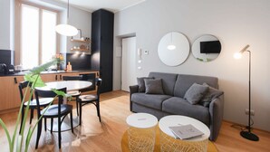 A modern living room with a grey sofa, a dining table with chairs, a kitchen area with a window, and a round mirror on the wall. - Cà Bèla Vitruvio - Flat 2 (Milano)