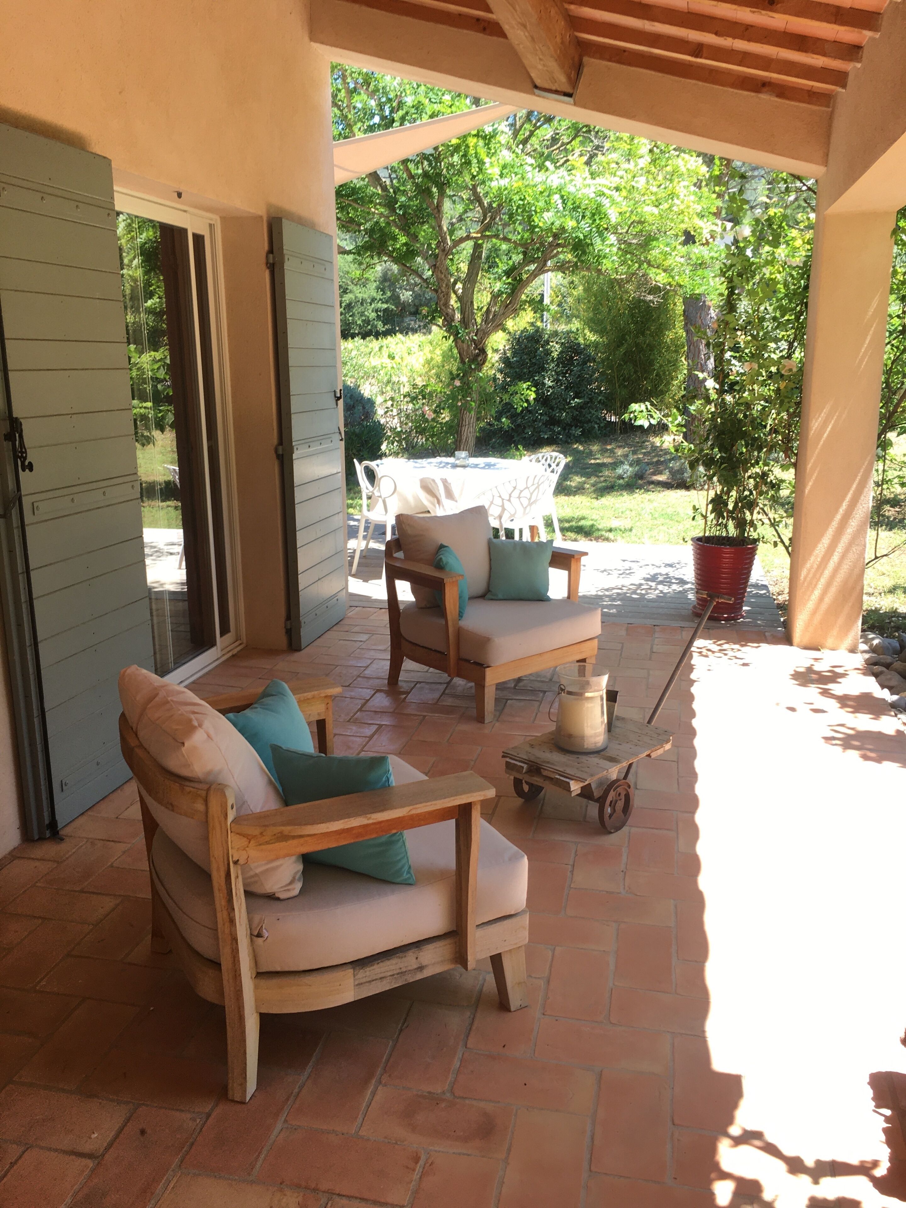 A covered patio with two wooden chairs and a small table, a potted plant, and a view of a garden with trees and a lawn.