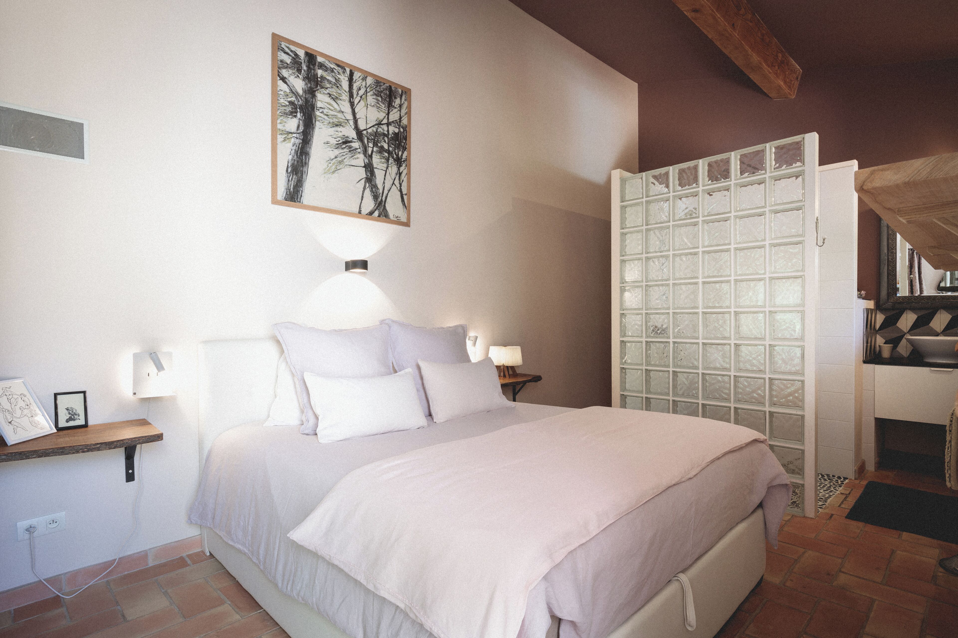 A bedroom with a bed, bedside lamps, a framed picture, a shelf with items, and a frosted glass door.
