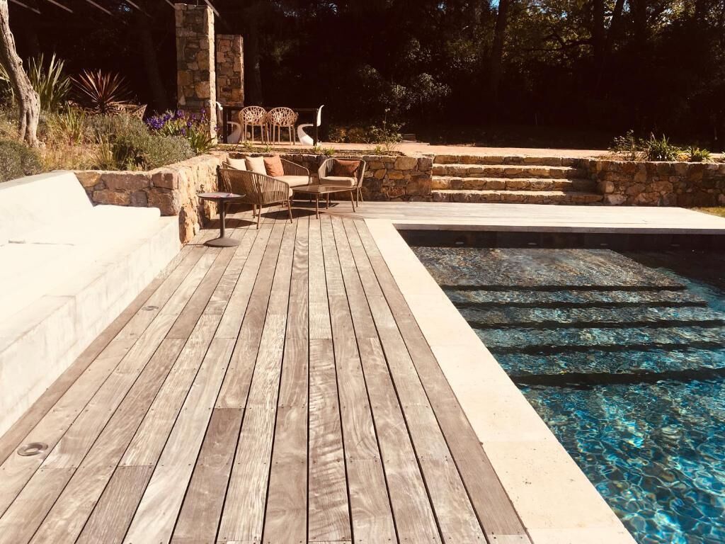 Una terraza de madera que conduce a una piscina con escalones de piedra y mobiliario exterior.