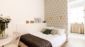 Une chambre à coucher comprenant un lit, une chaise, une table avec un vase de fleurs et un mur orné d’un motif géométrique et de tableaux encadrés.