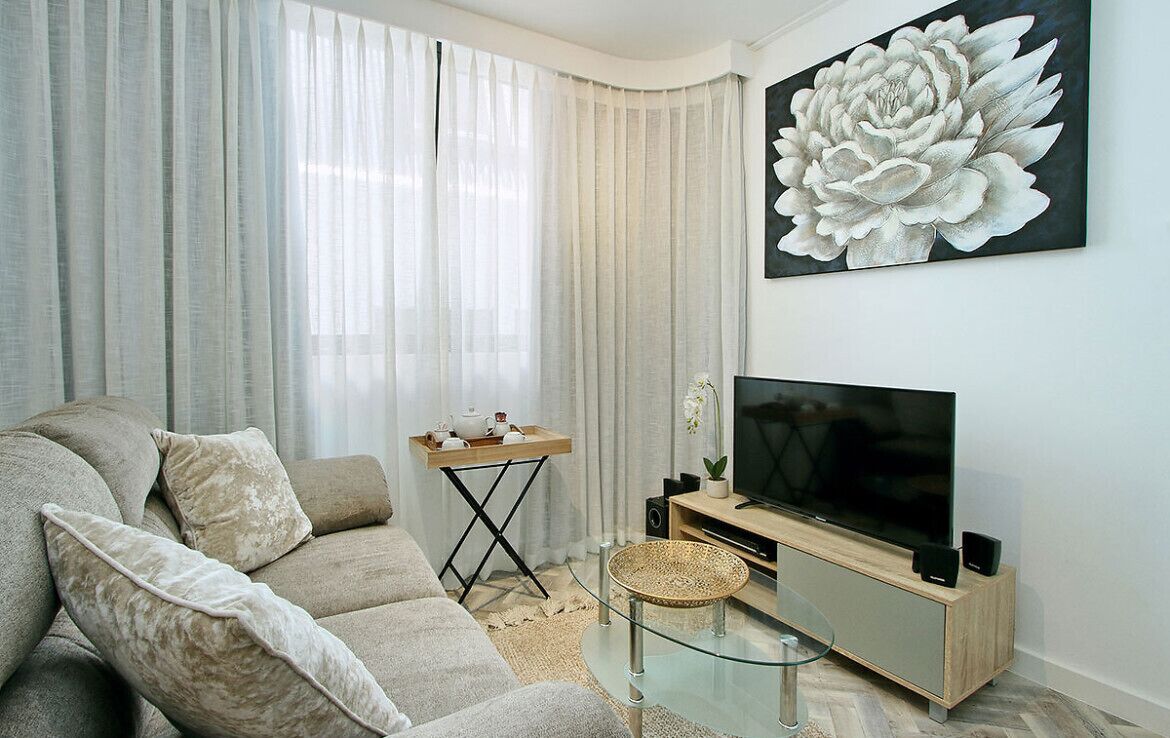 A modern living room with a beige sofa, a glass coffee table, a TV on a wooden stand, and a large floral painting on the wall.