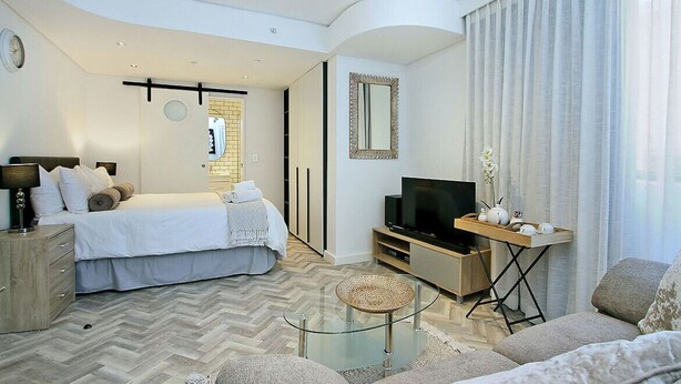 A modern hotel room with a bed, a TV, a sofa, a glass coffee table, and a small side table. - Tuynhuys (Cape Town)