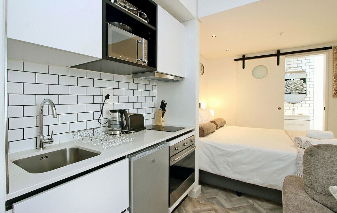 Uma cozinha compacta com azulejos brancos, uma pia de aço inoxidável e uma lava-louças pequena. Ao lado, um quarto com uma cama, uma mesa de cabeceira e um espelho.