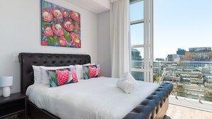 Moderni hotellihuone, jossa on suuri sänky, näköala kaupunkiin ja taulu seinällä.