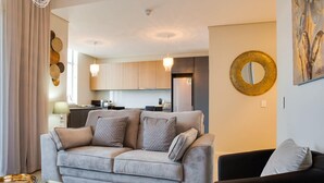 Una sala moderna con un sofá gris, un sillón negro y una mesa de centro de vidrio. Se ve una cocina al fondo con armarios de madera y un refrigerador de acero inoxidable.