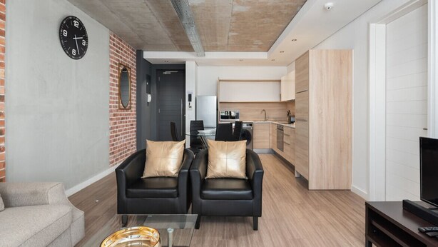 A modern living room with two black leather chairs, a glass coffee table, and a sofa. A kitchen area is visible in the background with wooden cabinets and a microwave. - Matrix (Cape Town)