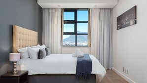 A bedroom with a large bed, a nightstand, a lamp, a window with curtains, and a cityscape view. - Matrix (Cape Town)