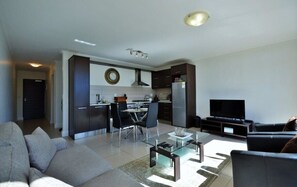 Uma sala de estar moderna com um sofá, uma mesa de centro e uma televisão. A área da cozinha é visível ao fundo, com armários e uma mesa de jantar.