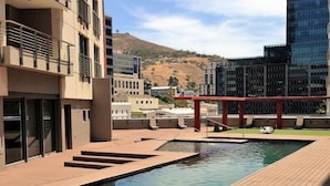 Un área de piscina moderna con cubierta de madera, una pérgola roja y vistas de edificios y colinas circundantes.