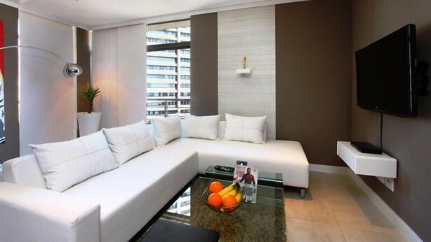 Una sala de estar moderna con un sofá seccional, una mesa de centro de vidrio con fruta, un televisor montado y vistas a la ciudad a través de amplios ventanales.