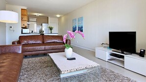 Ein modernes Wohnzimmer mit einer braunen Ledersofa, einem weißen Couchtisch mit einer Vase voller rosa Blumen, einem Flachbildfernseher auf einem weißen Standfuß und einer Küche im Hintergrund.